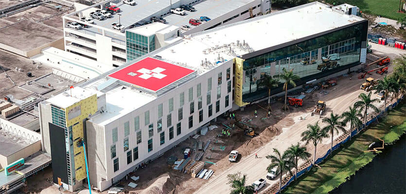 Aerial view of a hospital under construction featuring a red rooftop helipad with a white medical cross, surrounded by palm trees, construction equipment, and a nearby parking garage