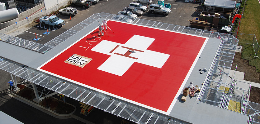 Workers applying fresh paint to a rooftop hospital helipad with a large white 'H' on a red landing surface